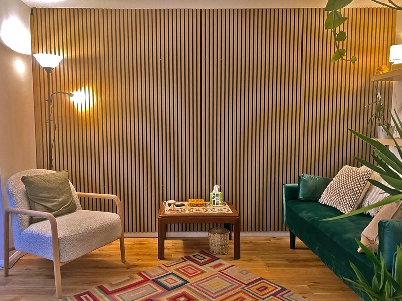 A side-on picture of the counselling space, facing the wall with the wooden slat panelling. On the left is a chair, on the right a green sofa, and in between is a 70's style coffee-table. To the left is a lamp casting a warm light across the wall